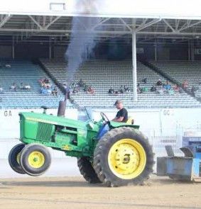 John Deere tractor pulling the sled in front of the grandstand, front wheels raised and exhaust smoke rising